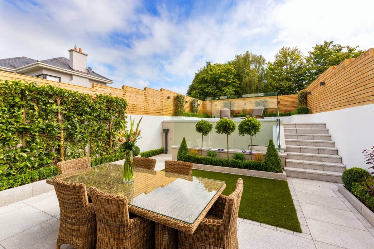 Multilevel backyard with wooden garden fence. Woven dining furniture with garden wall on lower level. Modern stone garden steps leading to lounge chairs