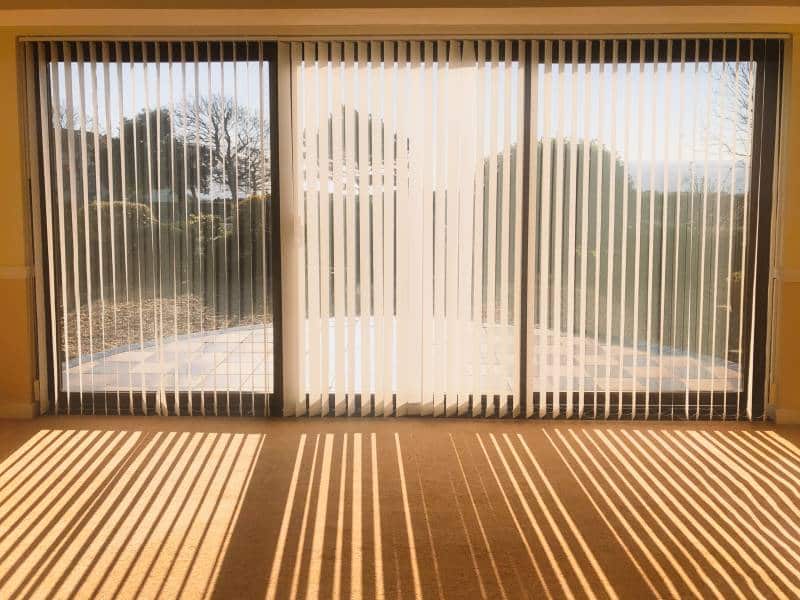 Sunlight shining through full length white vertical fabric blinds in front of three glass sliding french doors leading to a patio. Creating shadow stripes