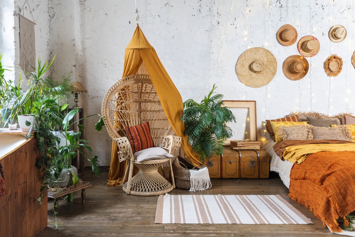 bohemian room with cozy interior, wicker chair, pillows, cushions, green plants in flower pot, bed and rug on wooden floor