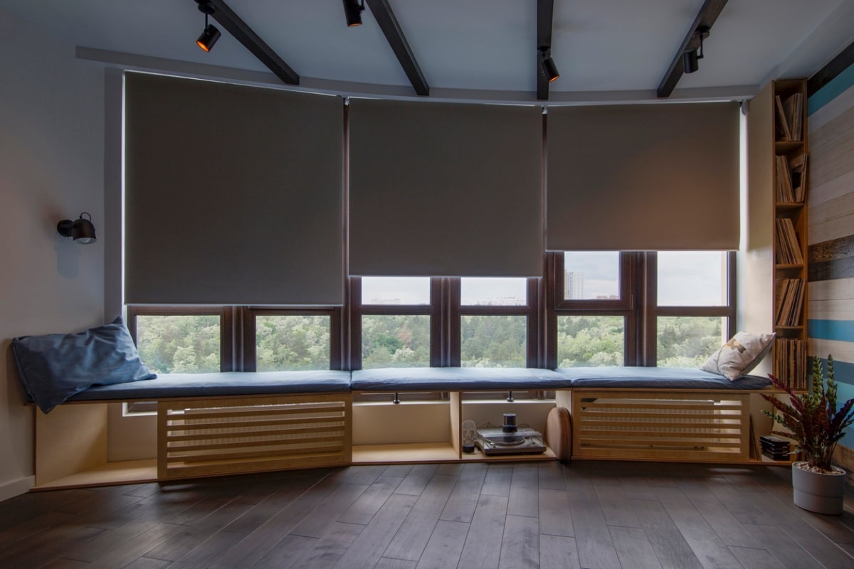 Automatic roller fabric blackout blinds on large windows above a windowsill with pillows