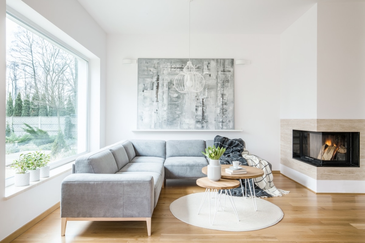 A living room with a gray sectional sofa, a coffee table, and a fireplace. There is a large painting on the wall and a window with a view of trees.