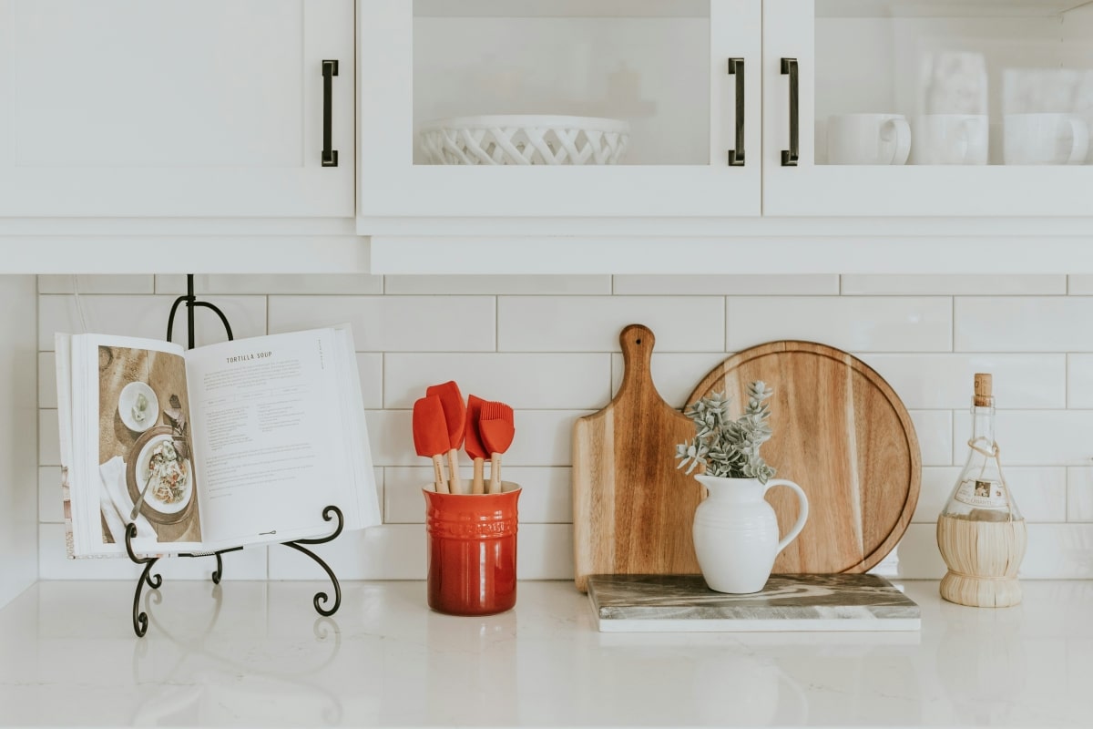 A white kitchen splashback with a cookbook on a stand, red spatulas in a jar, a cutting board, a vase with flowers, and a bottle of olive oil.