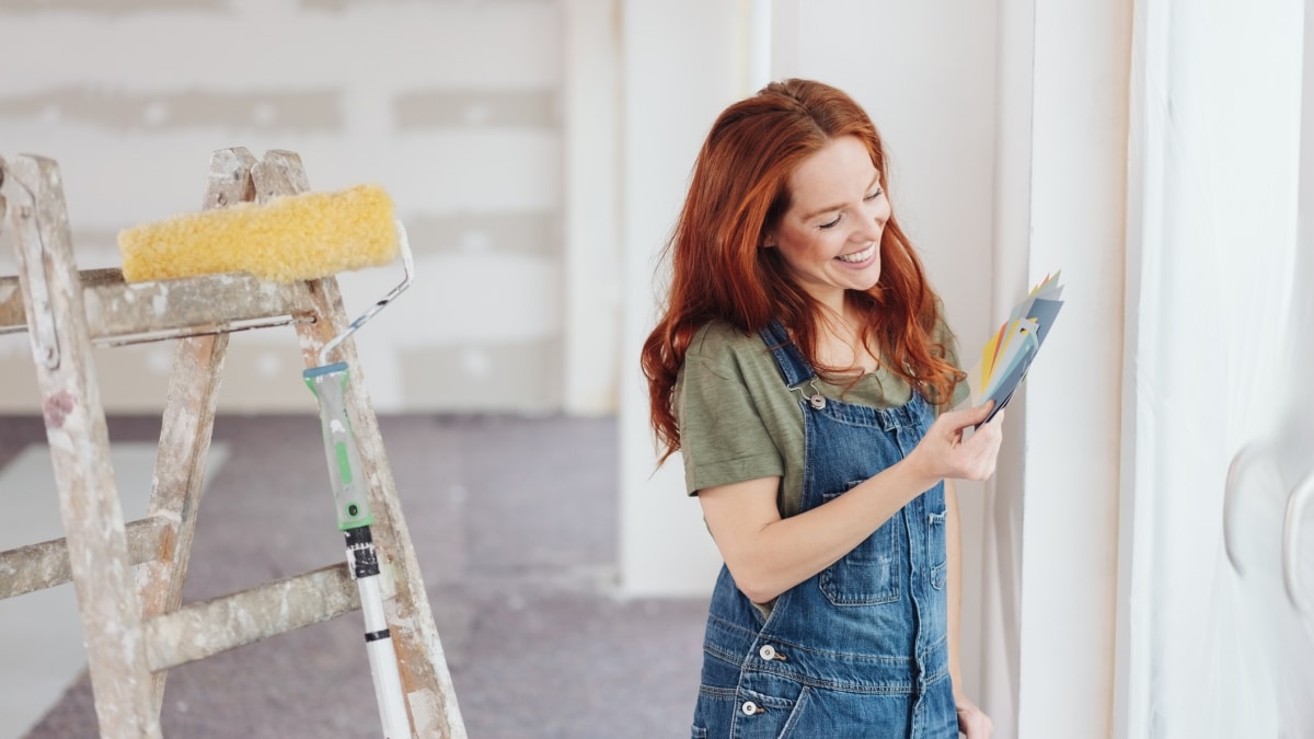 Smiling young woman comparing color swatches for the paint in her newly renovated unfinished living room with paint roller on the stepladder.