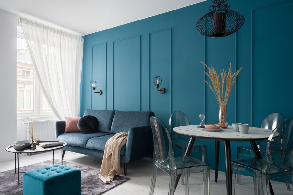 A modern living room with a blue accent wall, a gray couch, a round table with clear chairs, and a small table with a plant.
