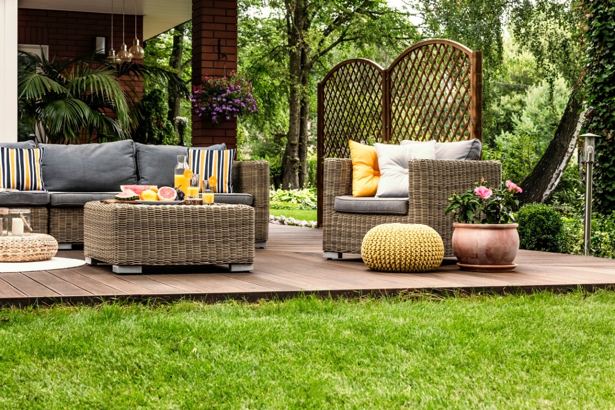 A wooden deck with a gray sofa, two chairs, and a table with drinks and fruit. There is a potted plant and a yellow pouf on the deck.