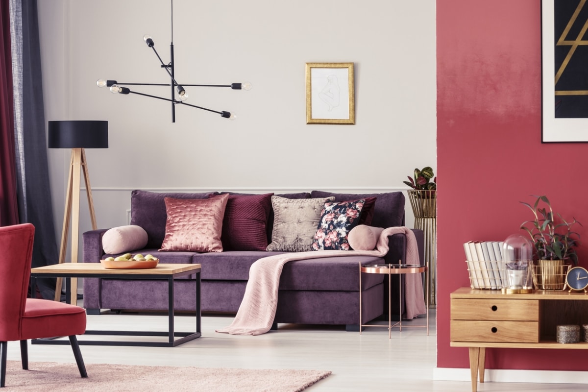 Modern living room with bold color accents, featuring a purple velvet sofa, pink and floral cushions, a red accent wall, and minimalist decor with gold and black details.