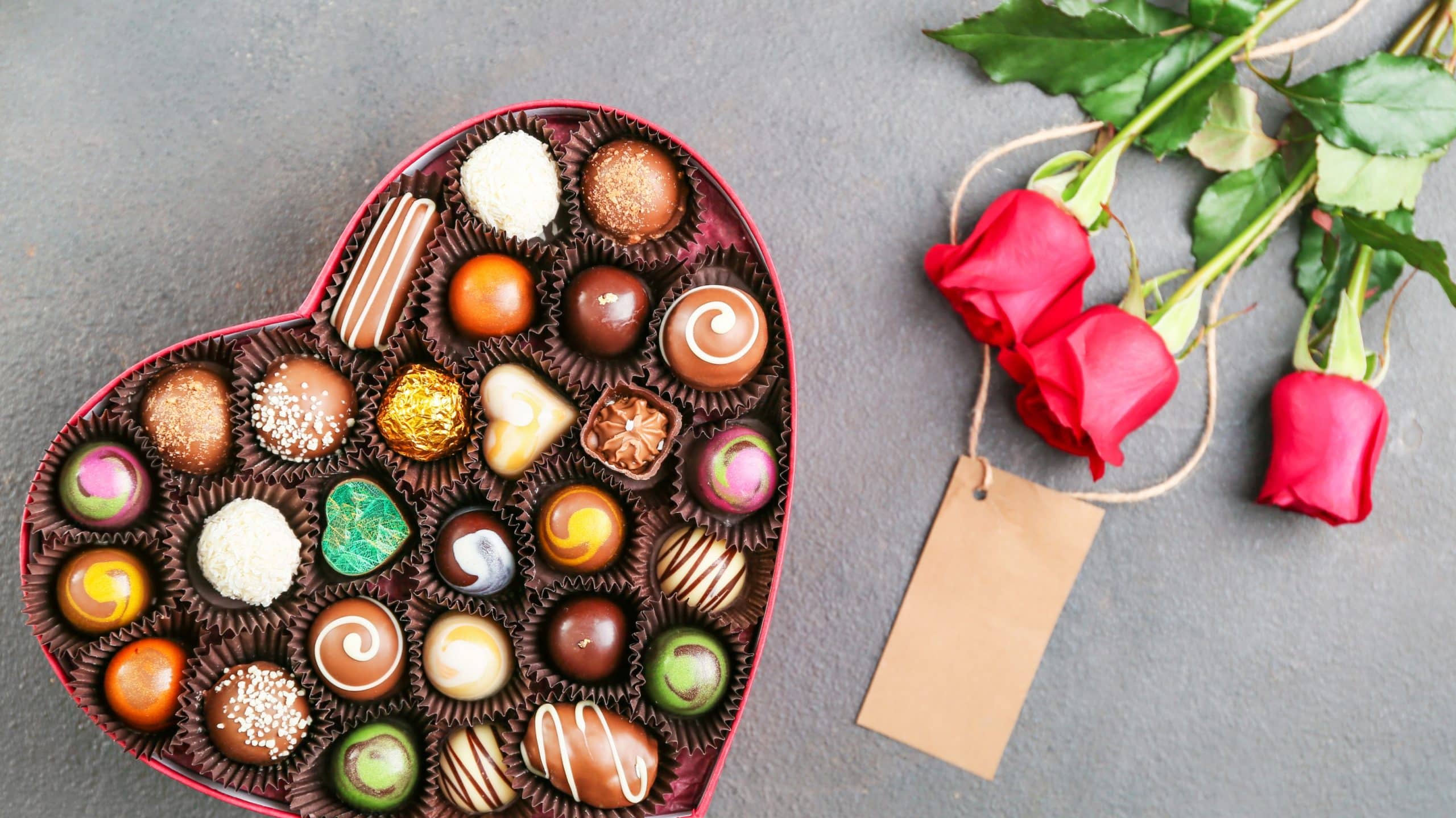 Heart-shaped box with delicious candies and bouquet of flowers on grey background