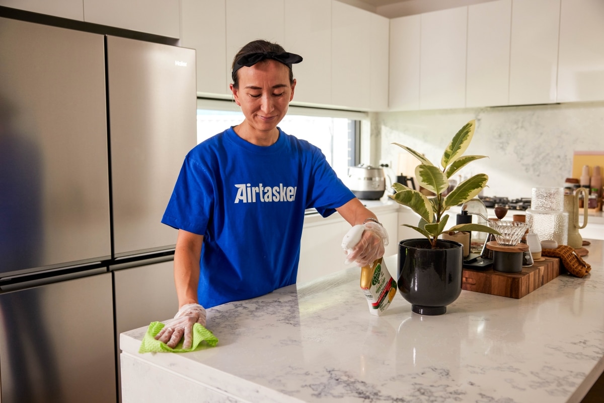 A Tasker in a blue Airtasker t-shirt cleaning a kitchen countertop with a disinfectant spray and cloth, ensuring a dust-free and allergen-reduced home.