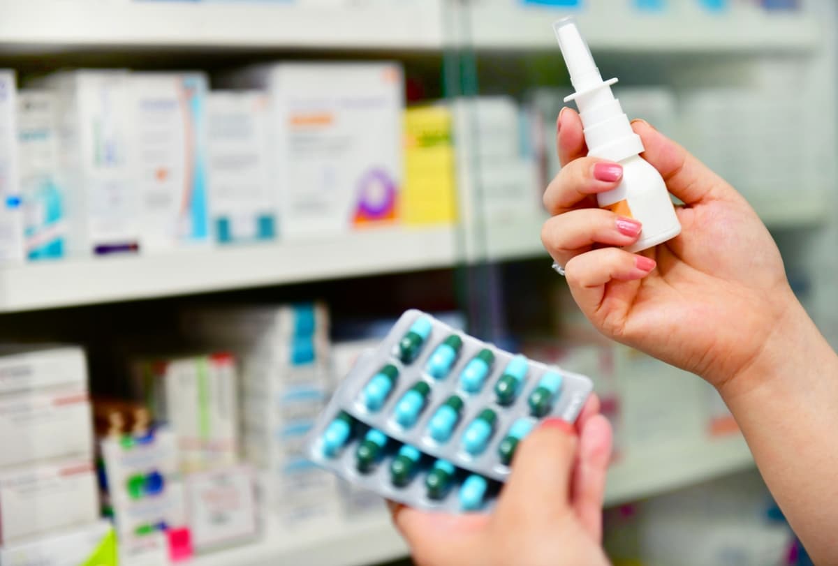 A woman's hands holding a white plastic nasal spray bottle and a blue blister pack containing green pills in a pharmacy aisle.
