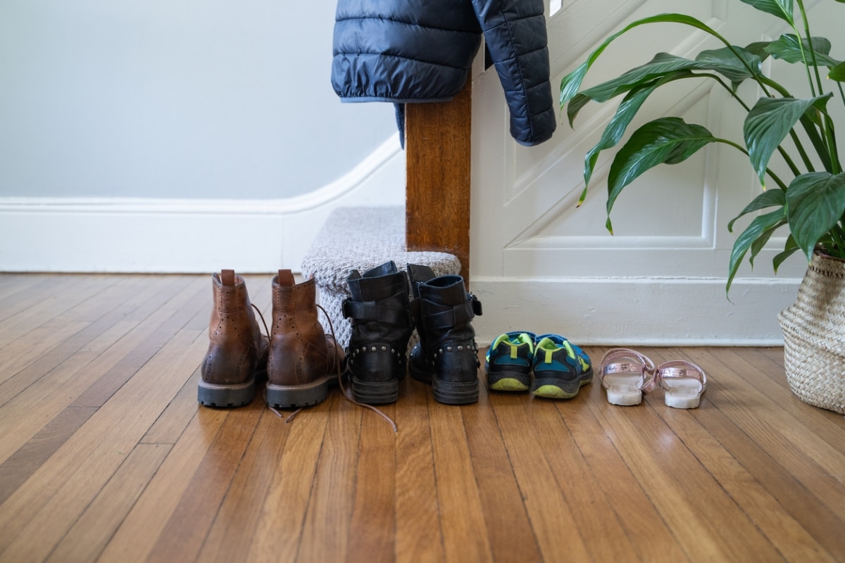 Image shows a hardwood floor with five pairs of shoes lined up in a row. Shoes include brown leather boots, black heeled boots, blue and green sneakers, and pink sandals. A dark blue puffer jacket hangs on a wooden post. A plant is on the right.