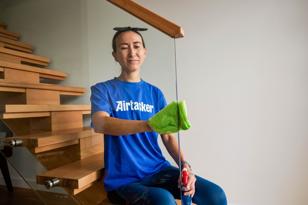 A Tasker wearing a blue Airtasker t-shirt wiping down a glass staircase railing with a microfiber cloth, removing dust and allergens to improve indoor air quality.