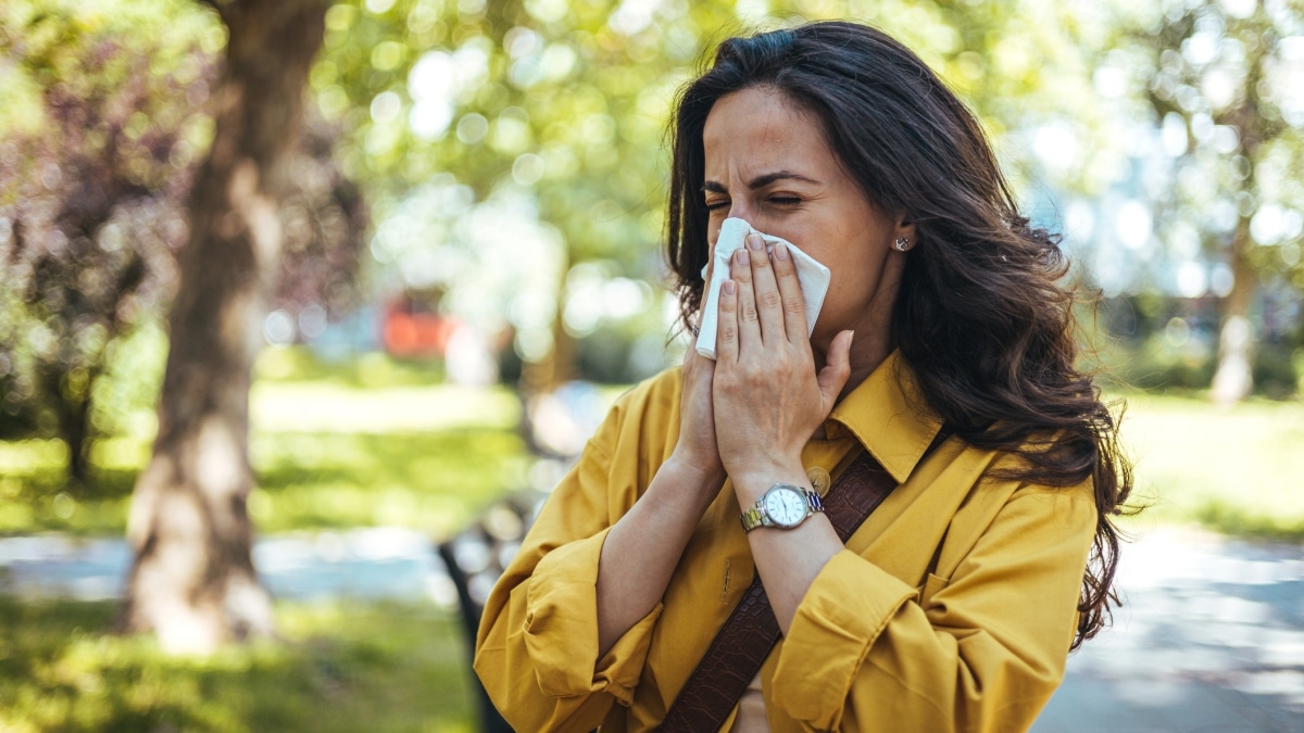 A woman in a mustard-yellow jacket sneezing into a tissue while outdoors in a park, surrounded by trees and greenery.