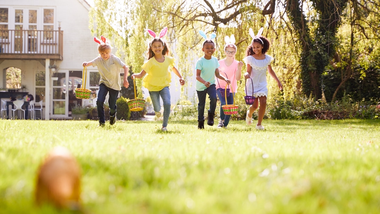 A group of children wearing bunny ears and running towards an Easter egg in their backyard.