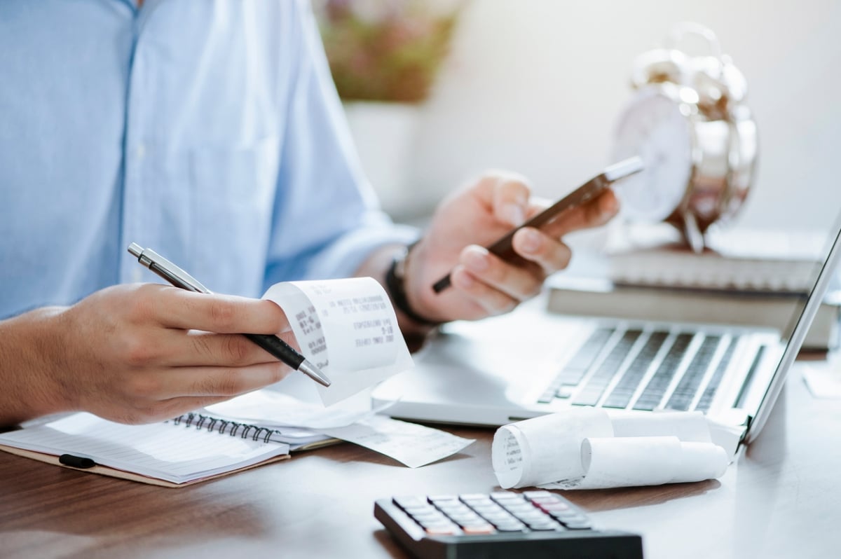 Individual holding a phone and receipts while sitting at a desk with a laptop and calculator, representing digital tax prep and filing tasks.