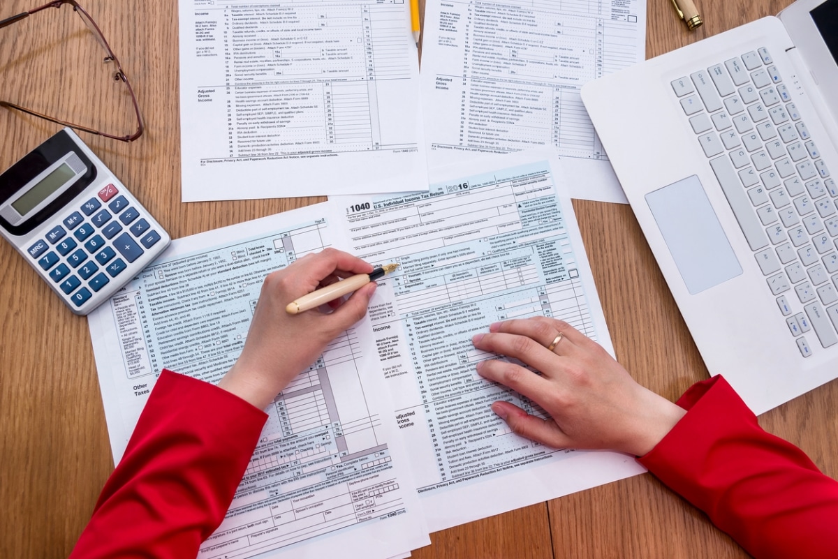 Close-up of hands filling out U.S. tax forms with a pen, calculator, and laptop on a wooden desk, illustrating detailed tax prep with official documents.