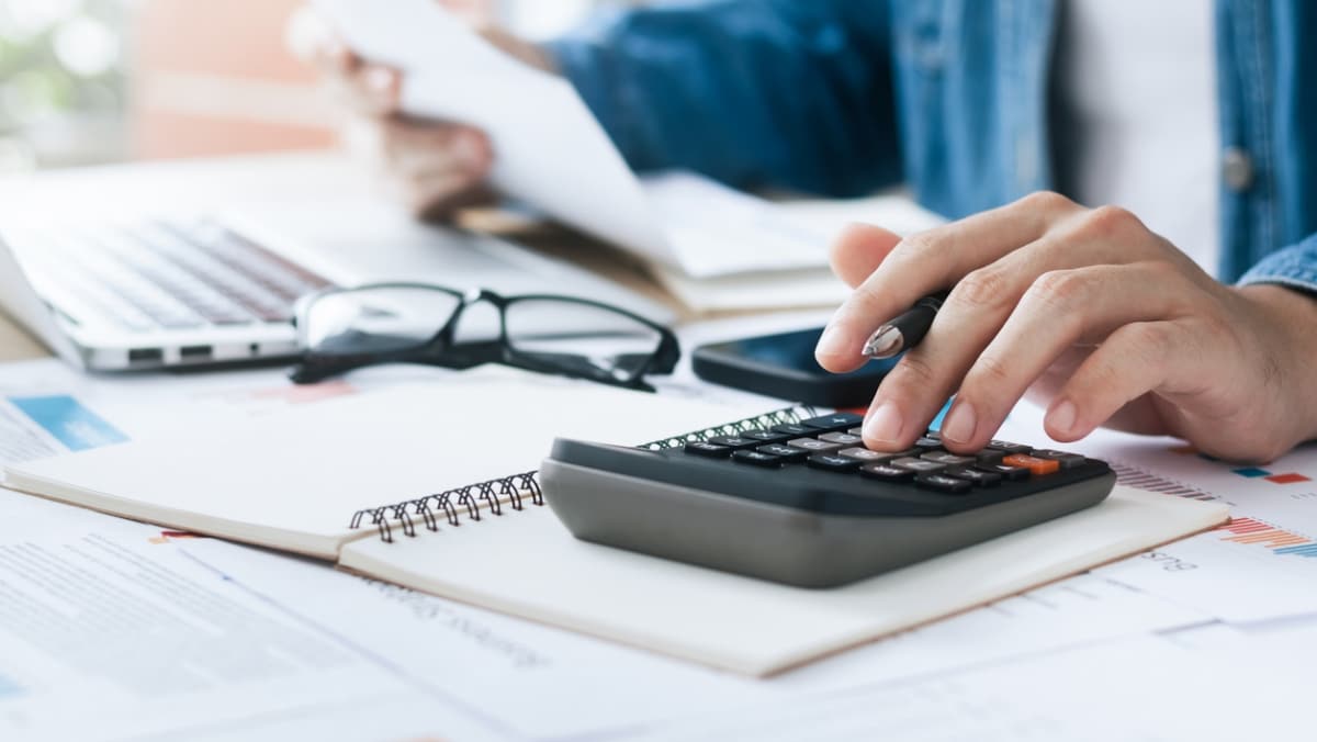 Person using a calculator and reviewing documents at a desk with a laptop and glasses, symbolizing tax prep and financial planning.